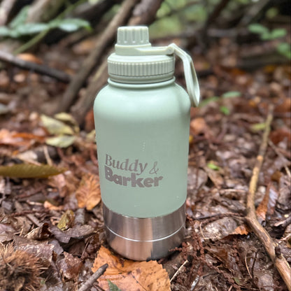 Sage green Trail Dog Travel Bottle with Buddy & Barker branding on a forest floor.
